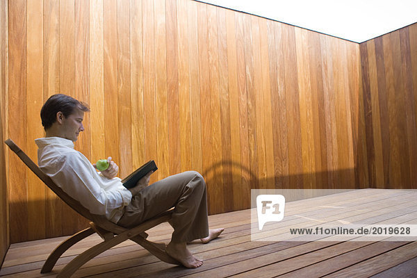 Man sitting in lounge chair  barefoot  reading book and eating apple  full length  side view