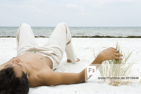 Junger Mann auf dem Rücken auf Sand liegend  Dünengras berührend  Ozean im Hintergrund