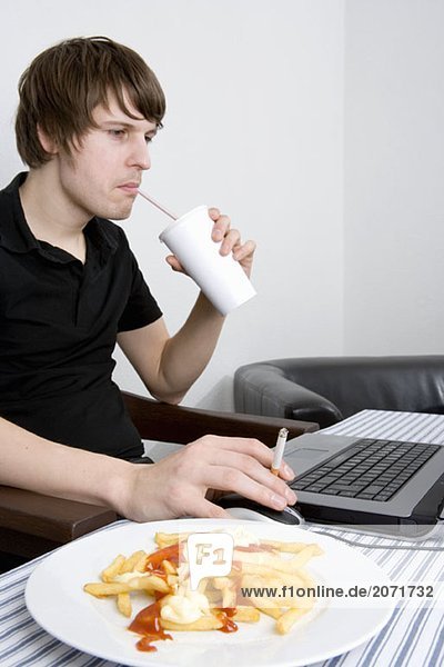 Ein junger Mann Zigarette rauchend am Computer neben dem ein Softdrink im Pappbecher und eine Portion Pommes-Frites stehen