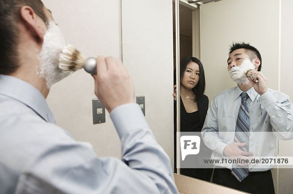 Ein Geschäftsmann steht mit Rasierschaum im Gesicht im Bad vor dem Spiegel  während er von einer hinter ihm stehenden Frau beobachtet wird