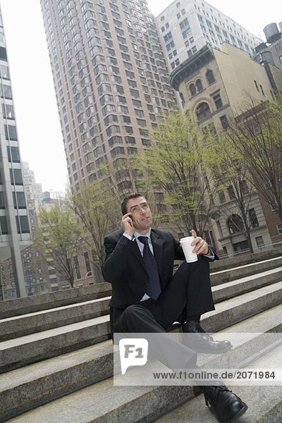 Ein Geschäftsmann sitzt auf einer Treppe und telefoniert einen Kaffeebecher haltend
