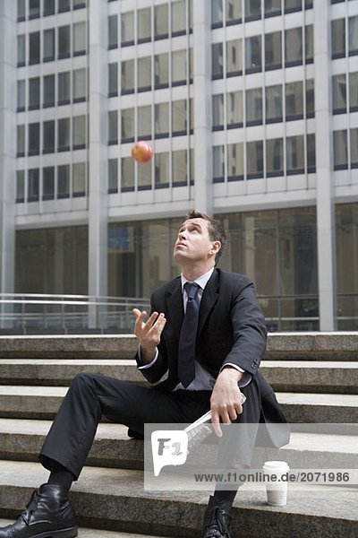 Ein Geschäftsmann sitzt während der Kaffeepause auf einer Treppe und wirft einen Apfel in die Luft