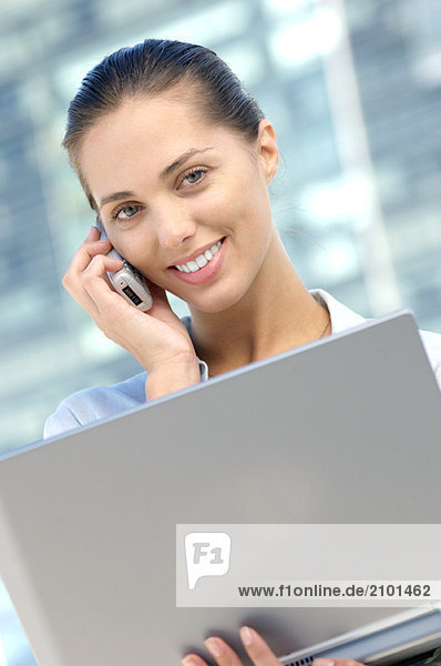 Businesswoman using mobile phone with laptop in hand  smiling