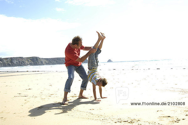 Boy doing handstand on beach with his father holding his legs