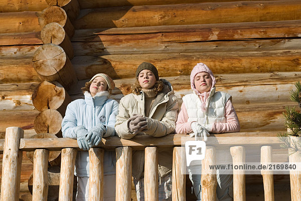 Three preteen or teen girls standing on deck of log cabin  eyes closed  low angle view