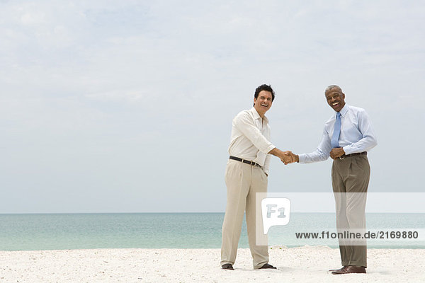 Zwei Geschäftsleute  die am Strand die Hand schütteln  beide lächeln in die Kamera.