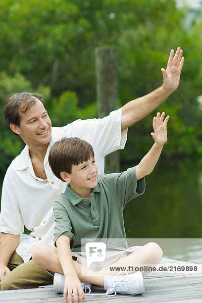 Father and son sitting on dock and waving together