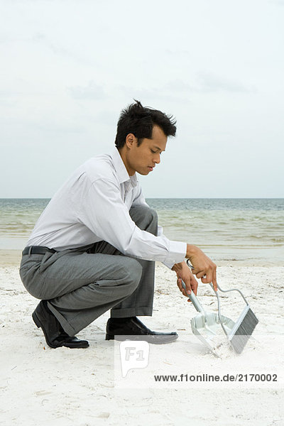 Mann hockt am Strand mit Besen und Kehrschaufel