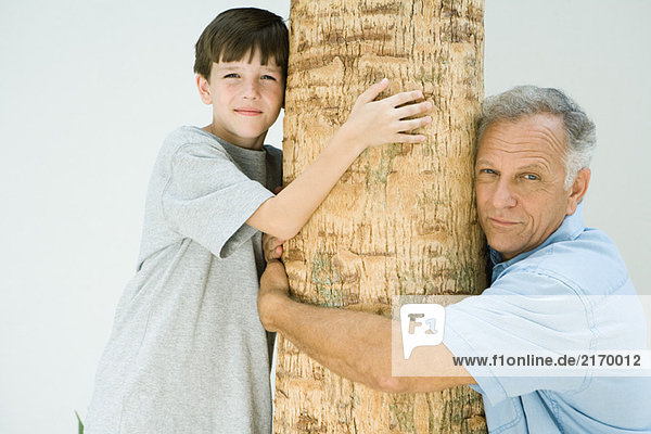 Großvater und Enkel umarmen Baumstamm  lächelnd vor der Kamera