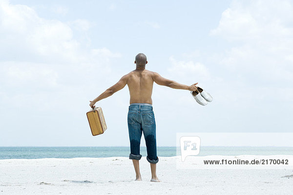 Man standing on beach holding out suitcase and shoes  rear view