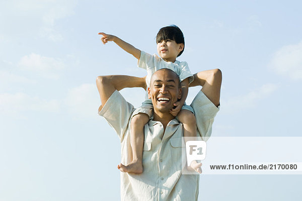 Father carrying son on shoulders and smiling at camera  child pointing and looking away