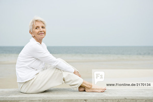 Seniorin am Strand sitzend  aufblickend und lächelnd