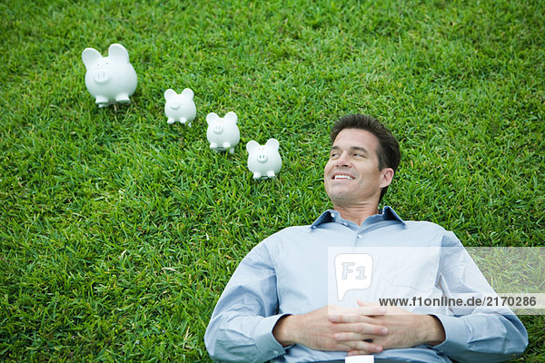 Mann auf Gras liegend  neben Sparschweinen  lächelnd