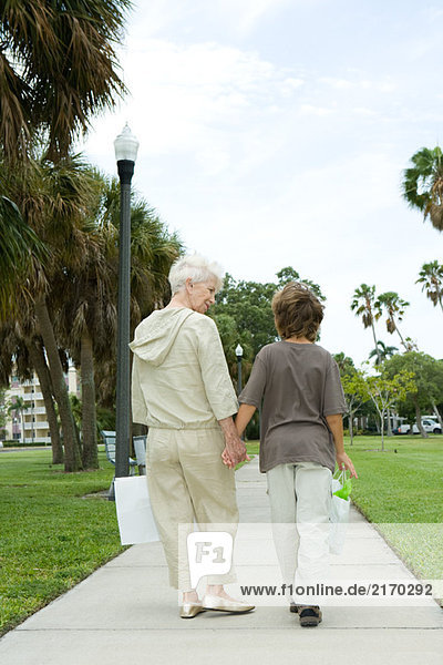 Junge und Großmutter gehen auf dem Bürgersteig  tragen Einkaufstaschen  reden  Rückansicht