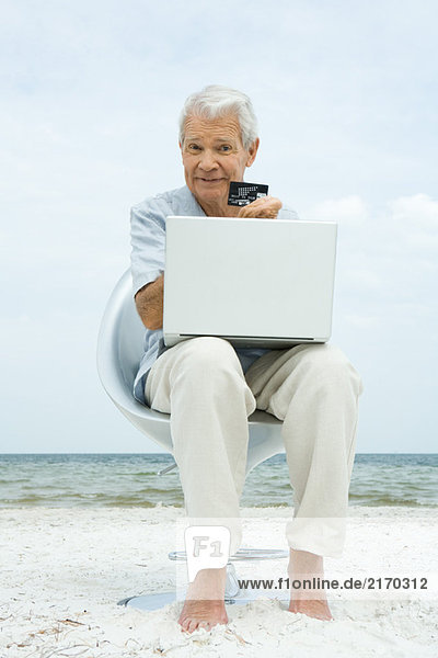 Senior Mann macht Kreditkartenkauf online  hält Kreditkarte  sitzt in Stuhl am Strand