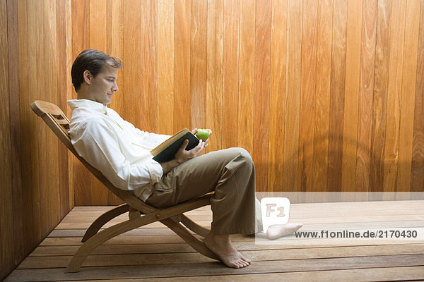 Man sitting in lounge chair reading book  apple in hand  side view