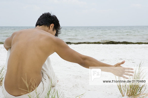 Mann sitzt am Strand und berührt Dünengras  Rückansicht