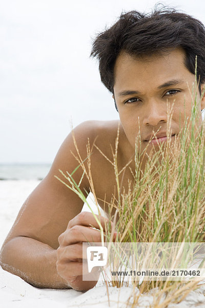 Mann liegt am Strand und berührt Dünengras  schaut in die Kamera.