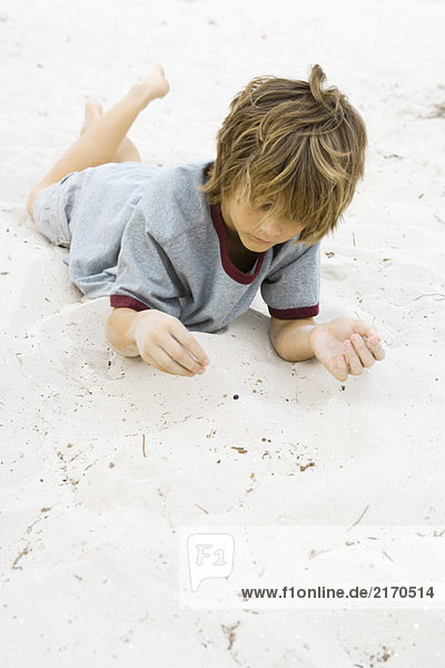 Junge auf dem Bauch liegend in Sand mit erhobenen Beinen  nach unten schauend