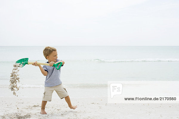 Kleiner Junge am Strand  der die Schaufel hochhält und Sand verschüttet.