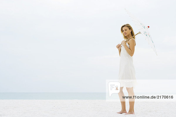 Frau steht am Strand und hält den Sonnenschirm  schaut weg.