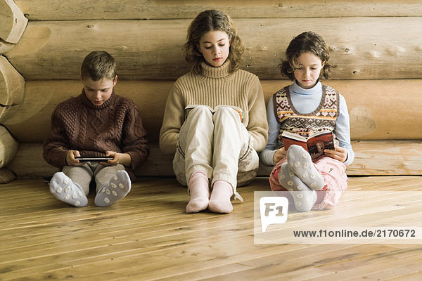 Drei junge Geschwister sitzen Seite an Seite  lesen und spielen Videospiel