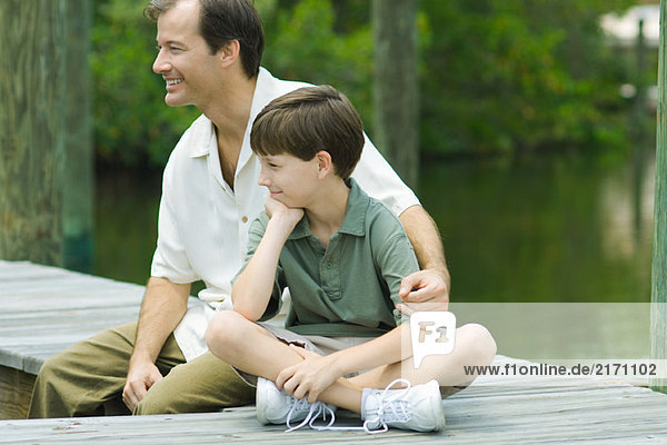 Mann sitzt auf dem Dock mit Arm um die Schulter des Sohnes  schaut weg und lächelt.