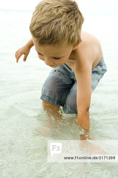 Kleiner Junge steht knietief im Meer  beugt sich vor  um Hand ins Wasser zu legen  Nahaufnahme