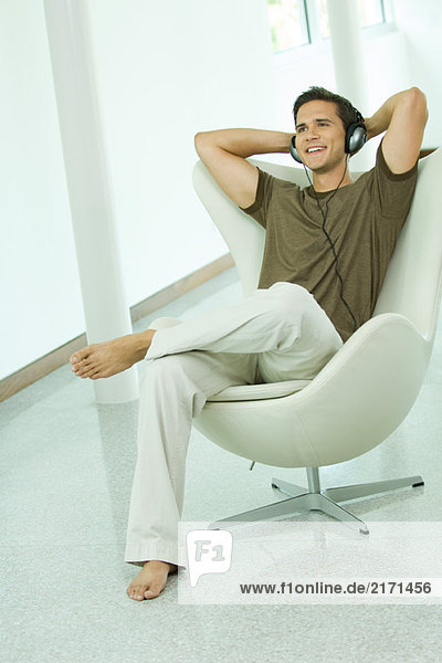 Young man listening to headphones with hands behind head  smiling  full length