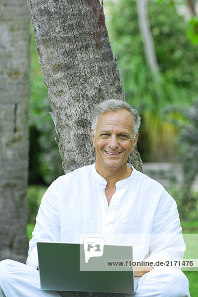 Mature man sitting  leaning against tree  using laptop computer  smiling at camera