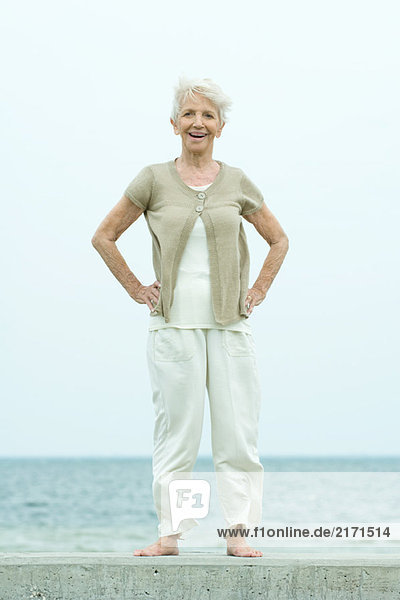 Seniorin stehend mit Händen auf den Hüften  lächelnd vor der Kamera  Ozean im Hintergrund  volle Länge