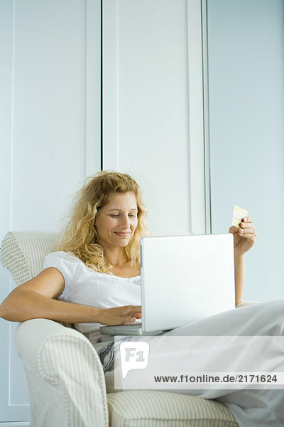 Woman making online purchase with credit card  sitting in armchair with laptop on lap
