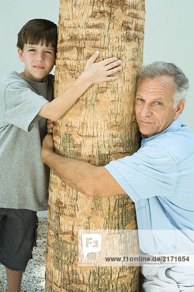 Grandfather and grandson hugging tree trunk  both looking at camera