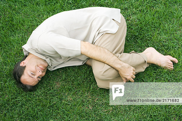 Mann liegt in fetaler Position auf Gras und lächelt vor der Kamera.