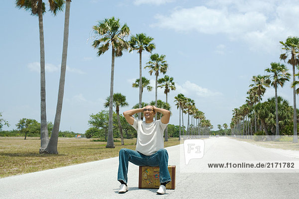 Man sitting on suitcase in the middle of street  hands on head  smiling