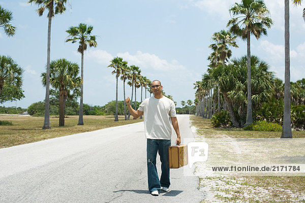 Man hitchhiking beside road  carrying suitcase