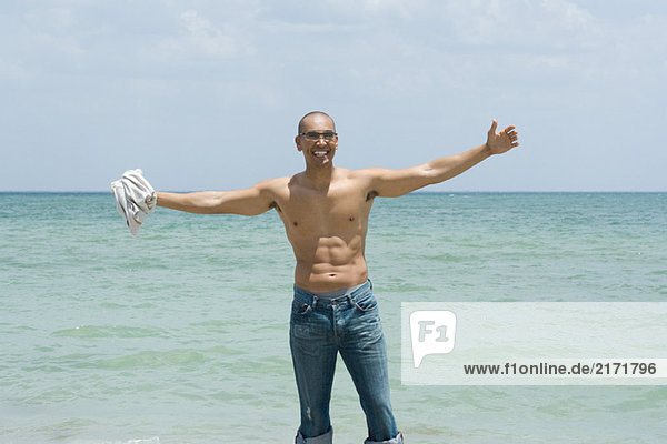 Man standing by the sea with arms outstretched  holding shirt in hand  smiling