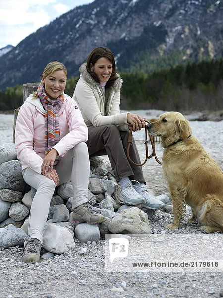 Zwei junge Frauen sitzen mit Hund am Flussufer