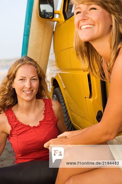 Zwei Frauen sitzen bei einem Van und lächeln mit Surfbrettern im Hintergrund.