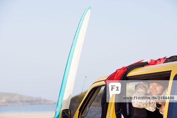 Mann im Van lächelnd mit Surfbrett an der Motorhaube lehnend.