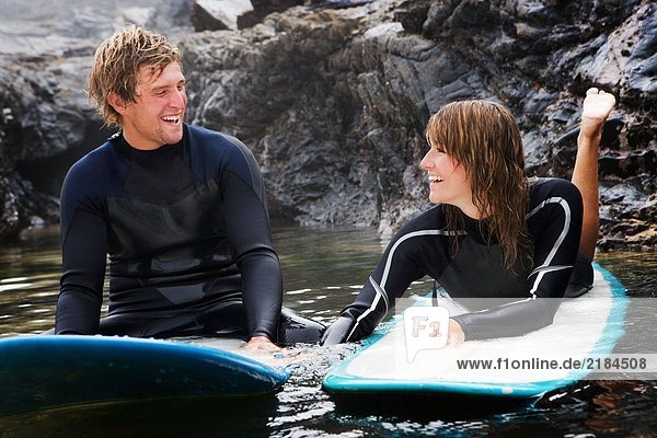 Ein Paar liegt auf Surfbrettern im Wasser und lacht.