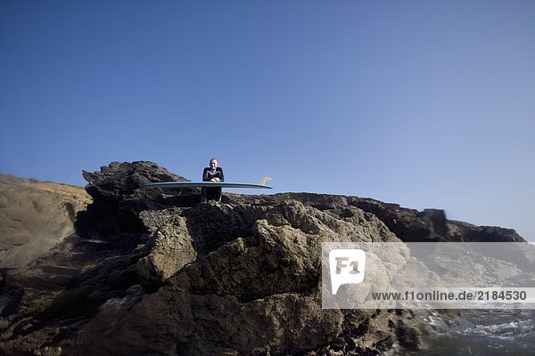 Mann sitzt mit Surfbrett auf großen Felsen und lächelt.