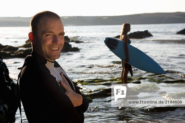 Mann steht lächelnd mit einer Frau  die das Surfbrett im Hintergrund hält.