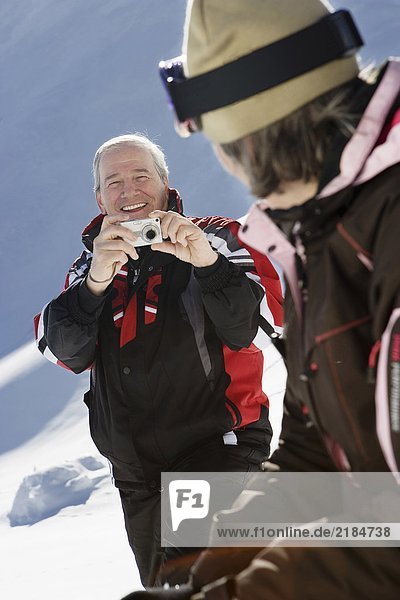 Mature man in ski-wear taking photograph of wife on mountain
