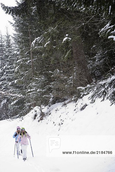 Zwei junge blonde Frauen  die mit Schneeschuhen den kleinen Hügel im Wald hinuntergehen.
