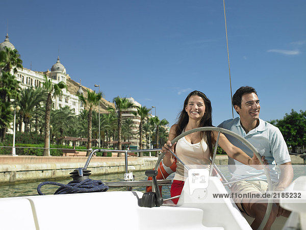 Young couple at wheel of yacht in marina  smiling