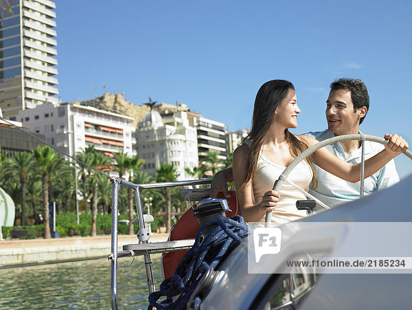 Young couple at wheel of yacht in marina  smiling