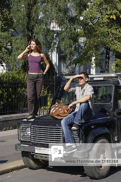 Frau steht auf der Motorhaube des SUV auf dem Handy  Mann sitzt auf der Motorhaube.