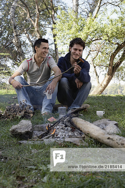 Two men cooking hot dogs over a fire pit smiling.