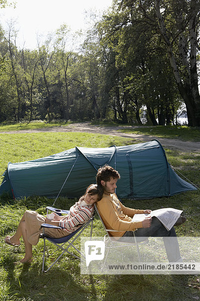 Ein Paar sitzt Rücken an Rücken auf dem Campingplatz mit dem Mann  der eine Zeitung liest.